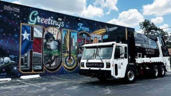 Garbage truck parked beside a colorful “Greetings from Houston” mural featuring Texas imagery, sports, and buildings under a bright sky.